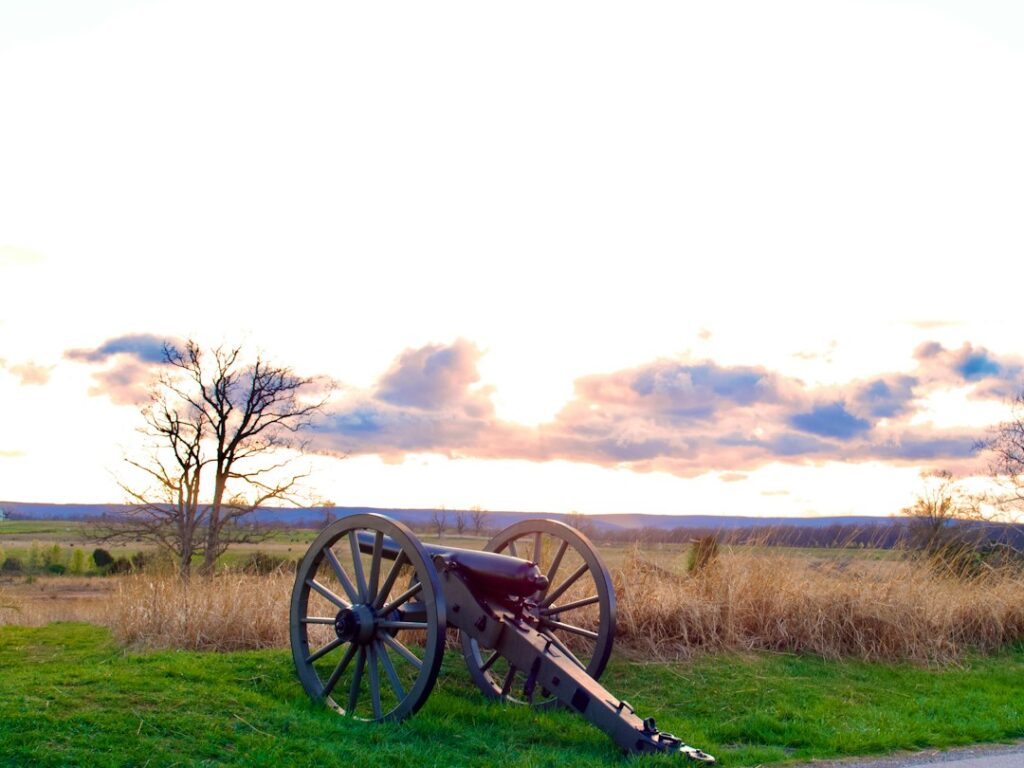 An old cannon sits on a grassy field with a bare tree under a bright sunset sky.