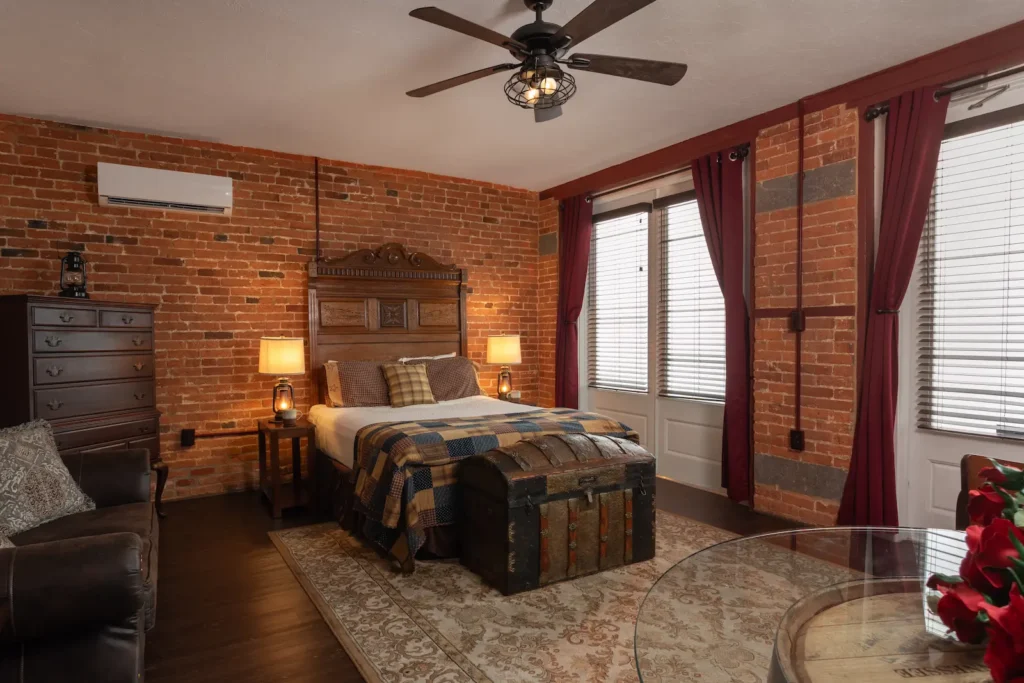 A bedroom with exposed brick walls, a wooden bed, a ceiling fan, and burgundy curtains.