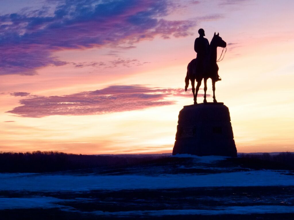 A silhouette of a person on a horse statue against a colorful sunset sky.