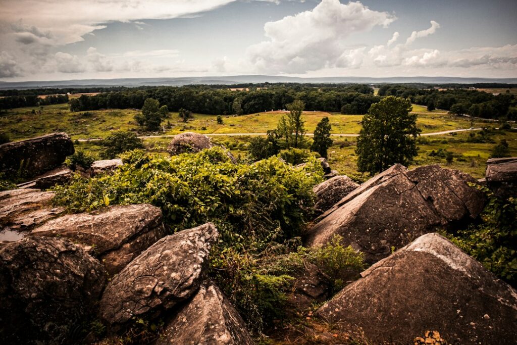 A rocky foreground overlooks a grassy field and distant trees under a cloudy sky.