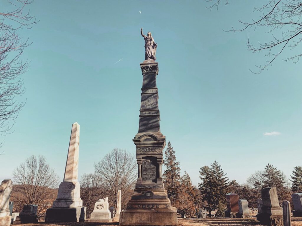 A tall, ornate stone monument with a statue on top stands in a cemetery under a clear blue sky.