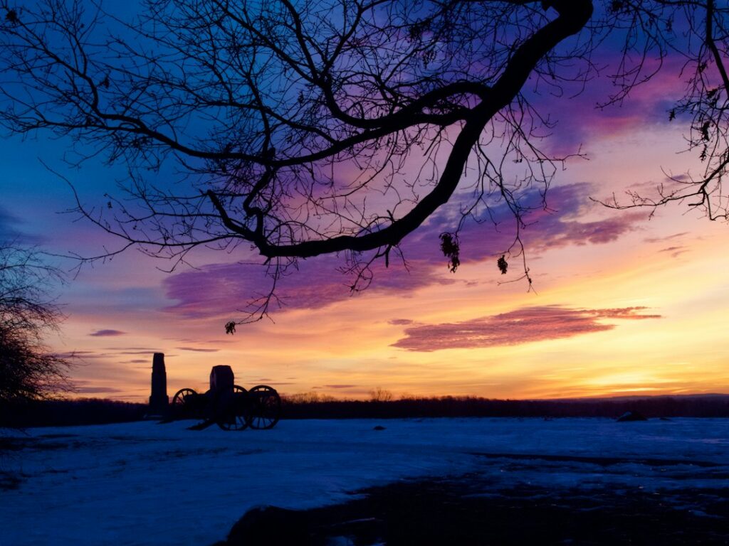 Silhouette of a cannon and monument under a vibrant, colorful sunset with a bare tree in foreground.